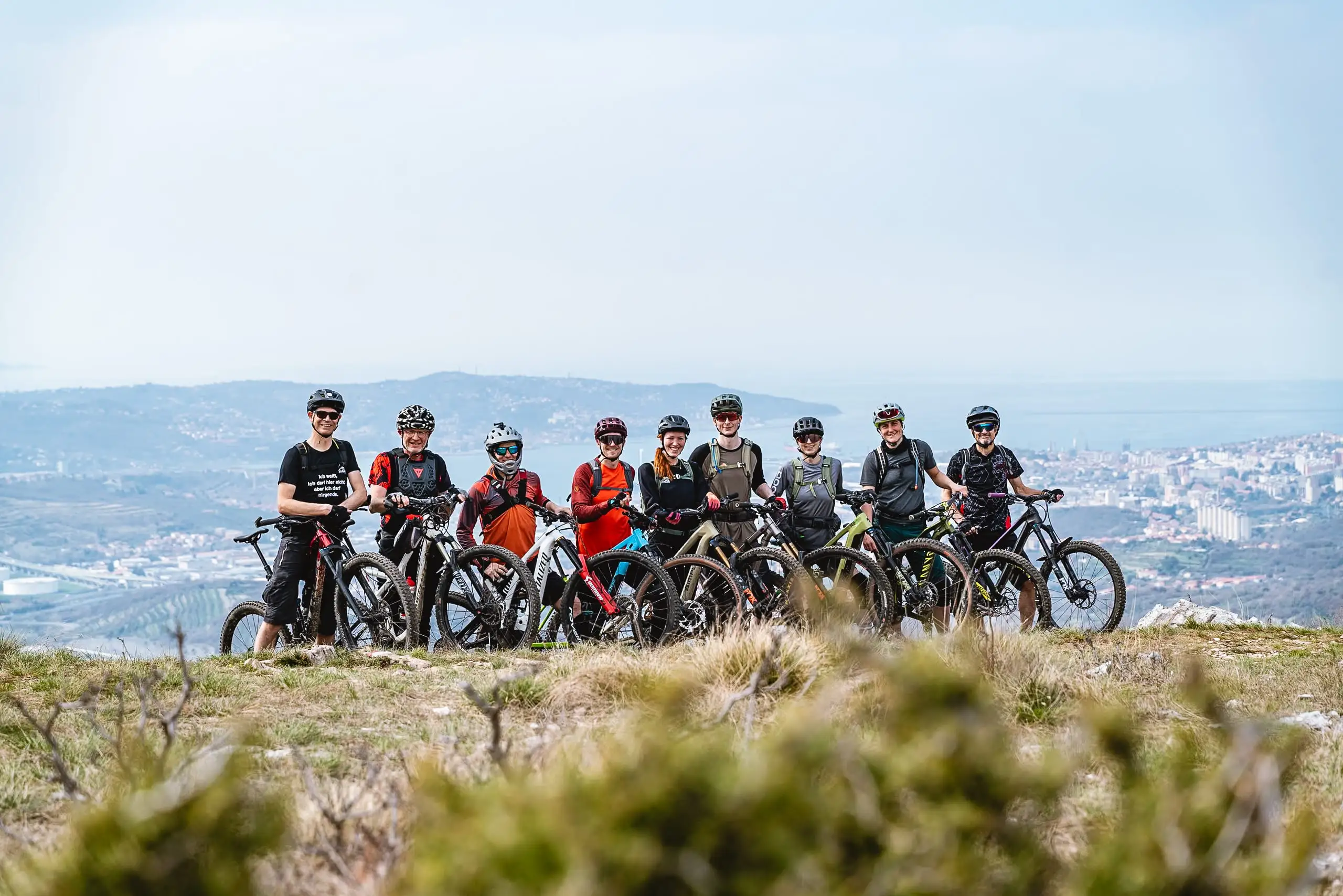 Eine Gruppe Mountainbiker:innen auf einem Hügel mit der Stadt Triest im Hintergrund.