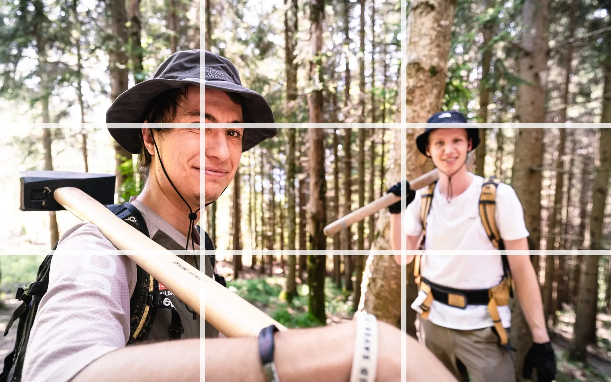 Zwei Trailbuilder stehen mit geschulterteten Werkzeugen im Wald. Als Overlay ist ein Raster gemäß der Drittel-Regel sichtbar. Einer der Schnittpunkte befindet sich am Auge des Motivs.