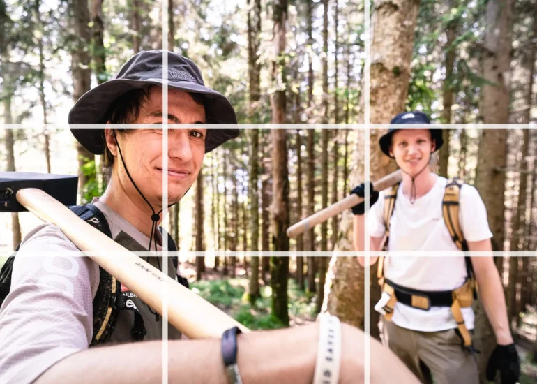 Zwei Trailbuilder stehen mit geschulterteten Werkzeugen im Wald. Als Overlay ist ein Raster gemäß der Drittel-Regel sichtbar. Einer der Schnittpunkte befindet sich am Auge des Motivs.