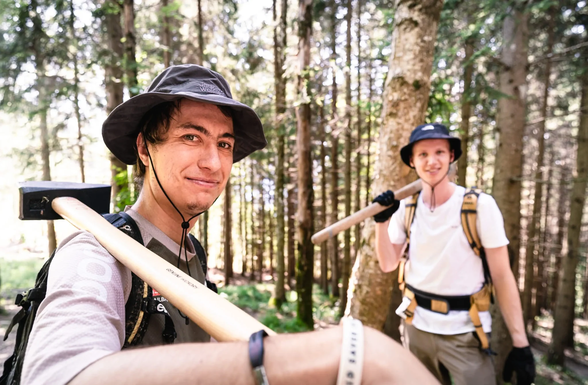 Zwei Trailbauer im Wald mit geschulterten Werkzeugen.